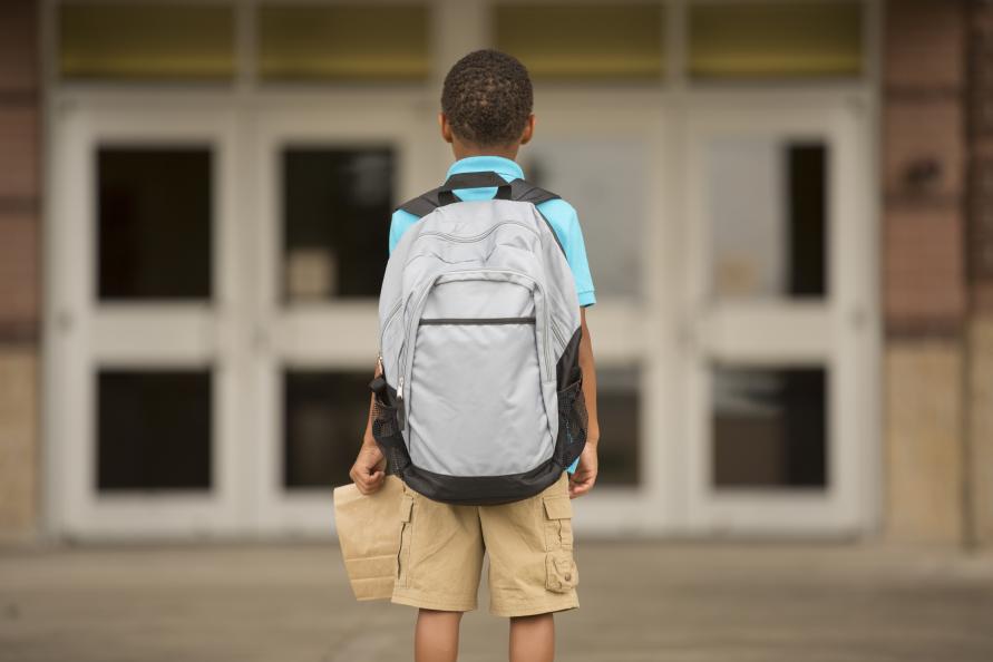 Pojke står framför skola med stor ryggsäck, fotograferad bakifrån.